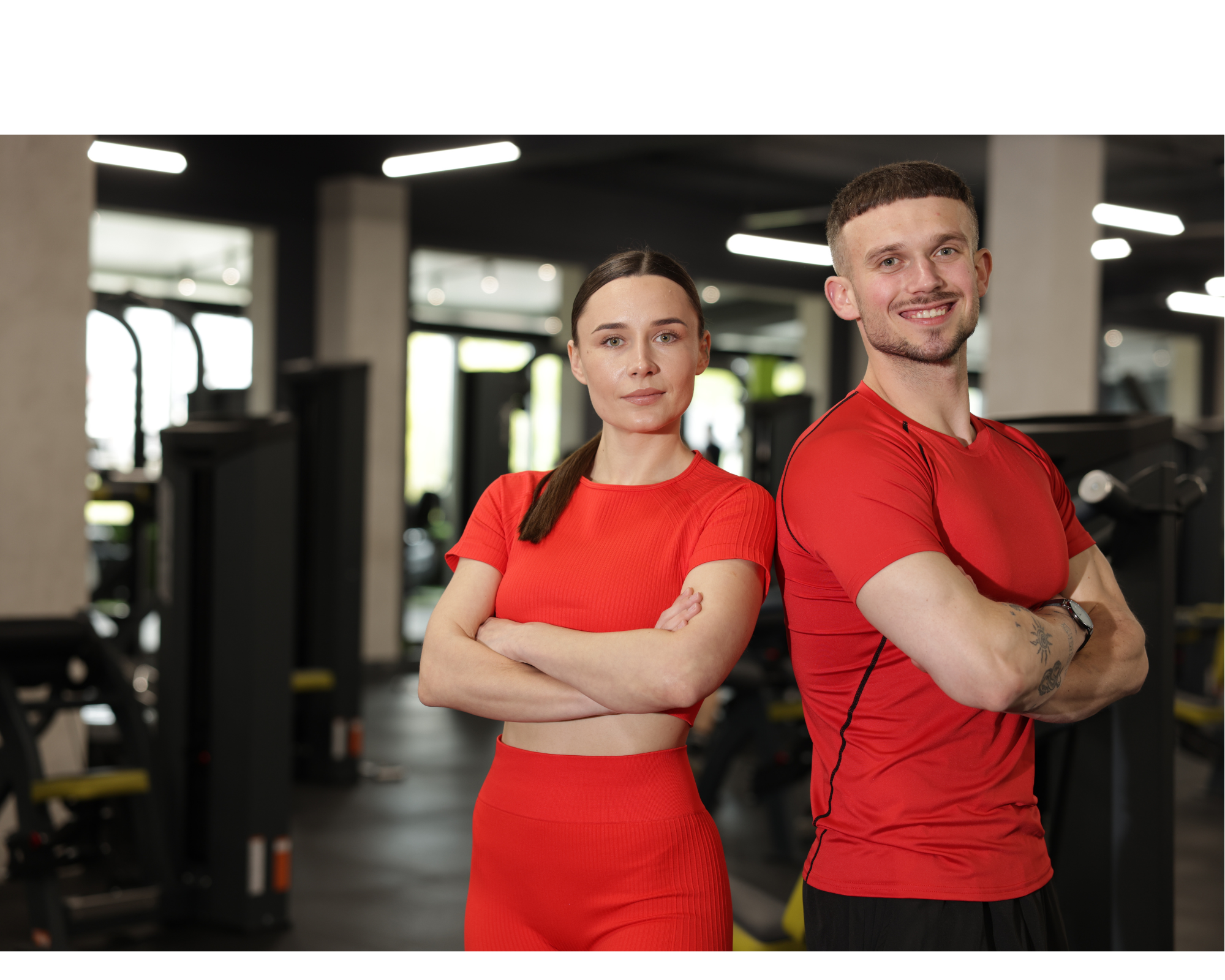 Two confident personal trainers standing side by side in a modern gym, both wearing red athletic wear, symbolising professionalism and strength in fitness coaching.