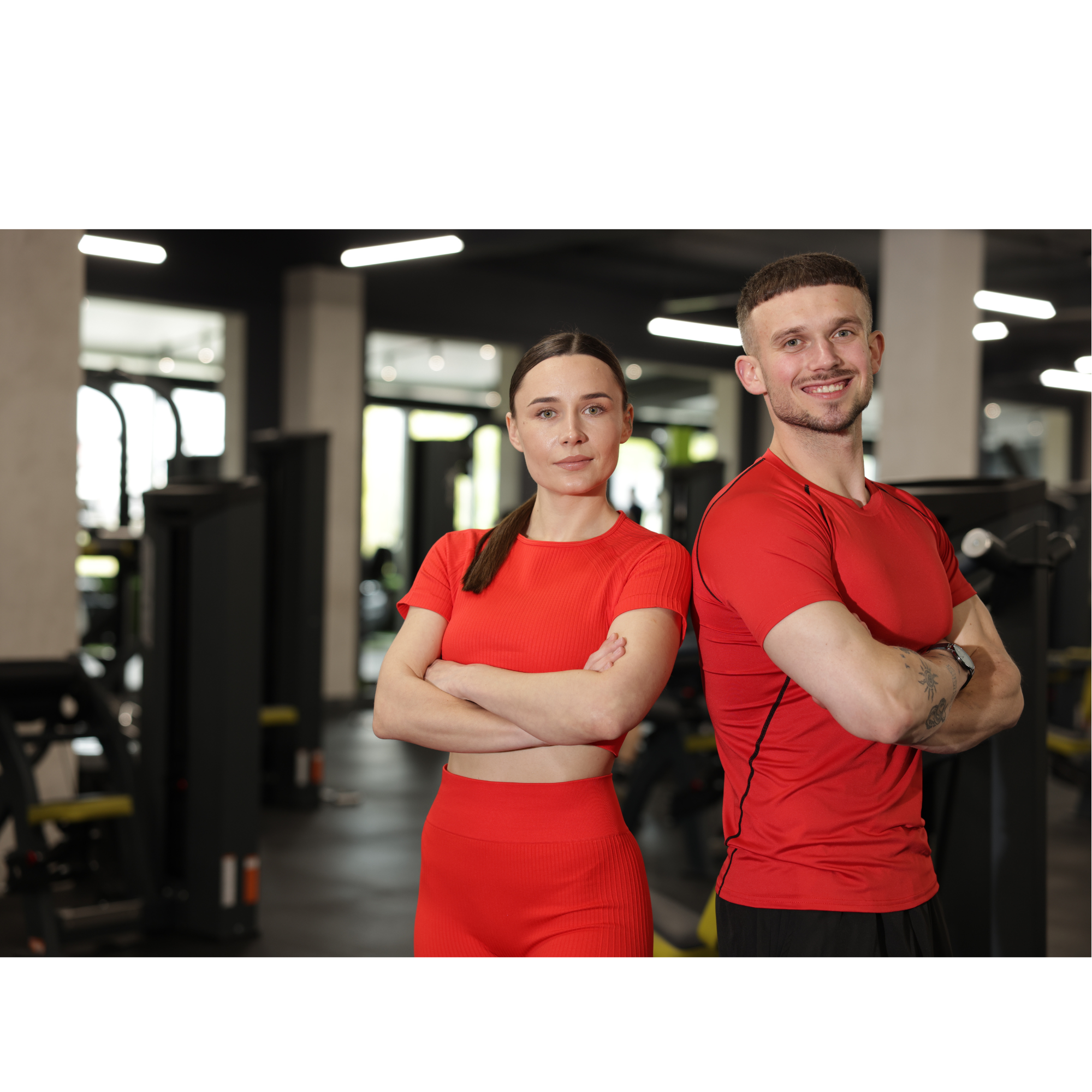 Two confident personal trainers standing side by side in a modern gym, both wearing red athletic wear, symbolising professionalism and strength in fitness coaching.